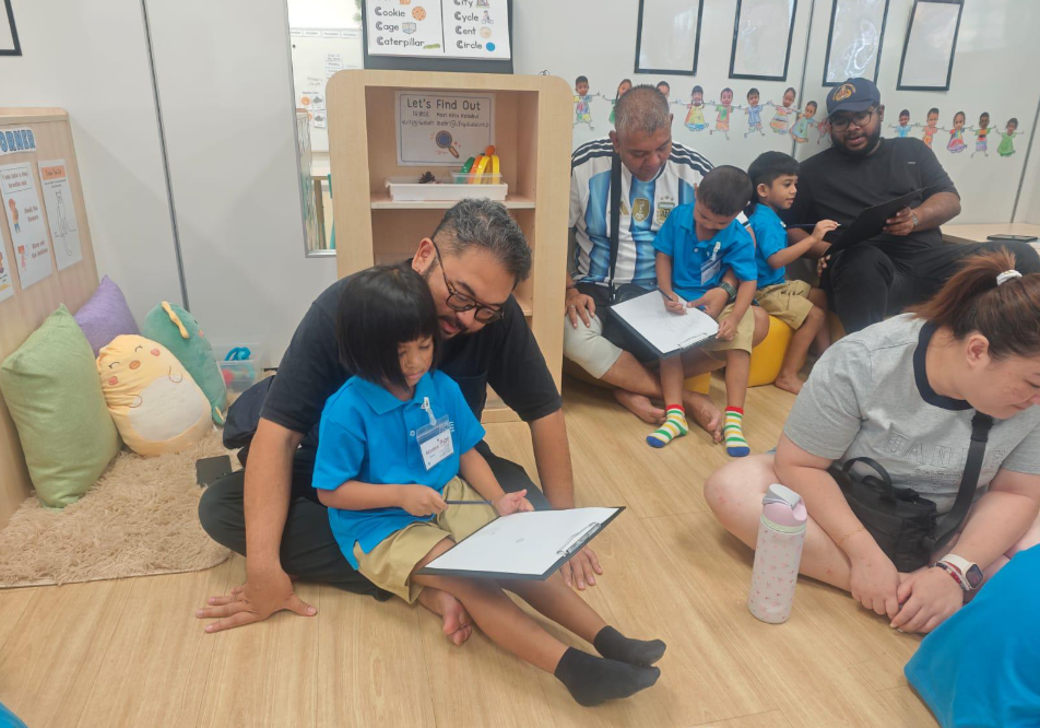 Adults sit with children in blue uniforms on a classroom floor, helping them write on clipboards.